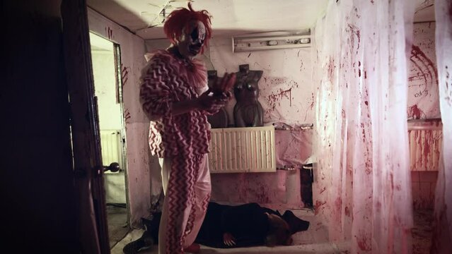 Psychopath maniac in a clown costume and his victim. The female body lies in a scary room with bloody streaks.