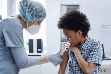 Female nurse in mask applying bandage on shoulder of little boy after vaccination at hospital