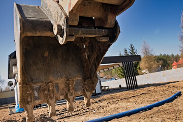 Łyżka kaparkowa z czterema stalowymi zębami , na końcu ramienia koparki , w trakcje kopania , na tle błękitnego nieba i nowego domu . Excavator working on a construction site. © Grzegorz