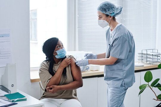 African young woman in mask talking to nurse while she applying plaster on her shoulder during procedure at hospital