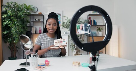 Experienced makeup artist of African American descent applies eye shadow on the eyelids, recording a tutorial on smoky eyes, in front of her on the table mirror, cosmetics and beauty accessories