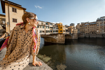 Fototapeta na wymiar Young woman enjoys beautiful view on famous Old bridge in Florence, sitting on the riverside at sunset. Female traveler visiting italian landmarks. Stylish woman wearing dress and colorful shawl