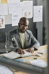 View of African young doctor sitting at table behind glass wall and examining ways of treatment in book