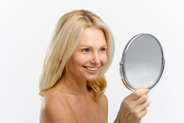 Cheerful blonde 50s woman examining her reflection in mirror isolated on white. Middle-aged female...