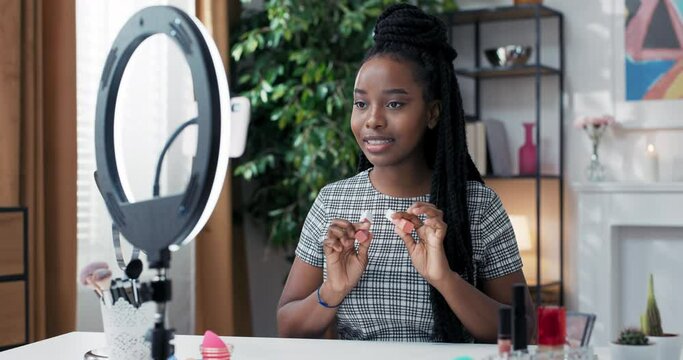 Female beauty blogger filming daily makeup routine tutorial using ring light with phone on tripod. Influencer with dark skin live streaming about cosmetics, comparing two shades of matte lipsticks.