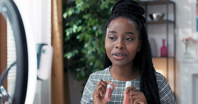 Young vlogger girl showing new kind of lipstick while recording vlog sitting at home. Woman record video indoors, advertising beauty products in front of phone camera.