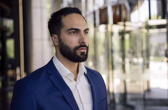 Young pensive Arabian business man wearing formal wear looking at window standing in modern office. Portrait of handsome Moroccan manager planning project at workplace. Successful business concept