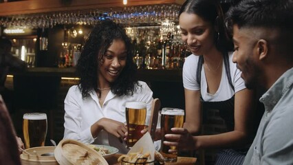 Multiracial waitress brings group of friends at restaurant their beers. The group of friends then clink their glasses.