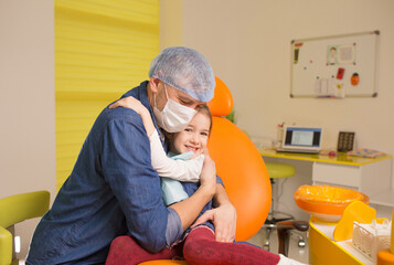 a male dentist hugs a little girl patient sitting in a dental chair