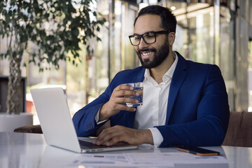 Handsome smiling Moroccan businessman using laptop computer working online sitting in modern office. Portrait of happy successful manager drinking water planning project sitting at workplace