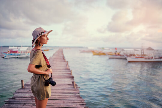 Photography and travel. Young woman in hat holding camera standing on wooden fishing pier with beautiful tropical sea view.