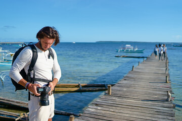 Photography and travel. Young man with rucksack holding camera standing on wooden fishing pier.