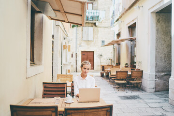 Obraz na płótnie Canvas Technology and travel. Working outdoors. Freelance concept. Pretty young woman using laptop in sidewalk cafe on ancient europian street.