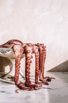 An octopus in an old and decorated footed dish, the tentacles falling outside on a marble table.