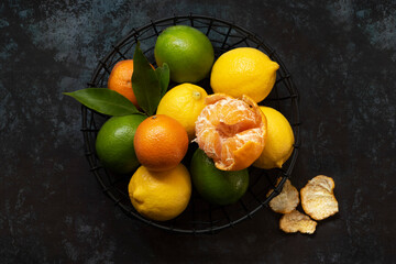 Lemons, limes and mandarins in a wire fruit basket. One mandarin is partially peeled.