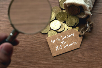 A hand holding a magnifying glass pointing to a bag of gold coins with a tag written with Gross Income vs Net Income. - obrazy, fototapety, plakaty