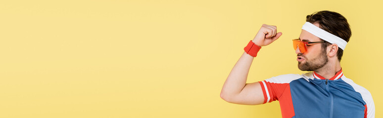 Young sportsman in sunglasses looking at muscles isolated on yellow, banner.