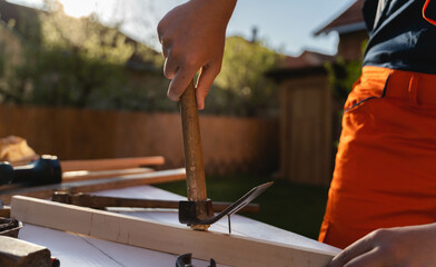 A man is hammering a nail in a plank on his workbench during the day