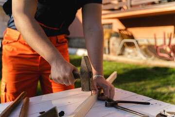 A man is hammering a nail in a plank on his workbench during the day