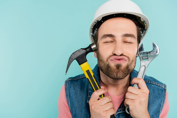 Young man in hardhat pouting lips and holding tools isolated on blue.