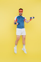 Sportsman in sunglasses jumping and holding dumbbells isolated on yellow.