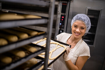 Portrait of female baker in white uniform and hairnet working in bakery production moving tray with...