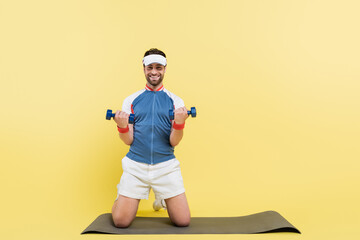 Cheerful sportsman holding dumbbells and looking at camera on fitness mat on yellow background.
