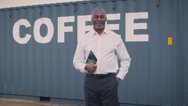Smiling mature male manager holding digital tablet at freight haulage business standing outdoors in front of shipping container labelled coffee looking into camera -shot in slow motion