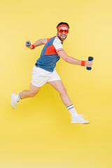 Fototapeta na wymiar Smiling sportsman in sunglasses jumping and holding dumbbells isolated on yellow.