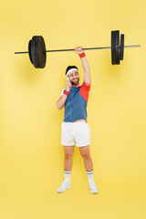 Full length of smiling sportsman talking on cellphone and lifting barbell on yellow background.