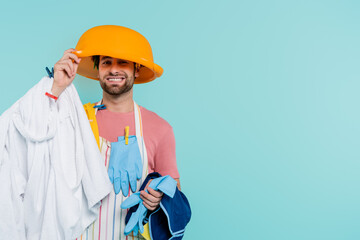 Cheerful man in clothespins holding wash bowl on head and clothes isolated on blue.