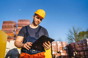 One man construction worker standing at warehouse or on the site holding clip document checking supply building material wearing protective helmet making inspection and control copy space