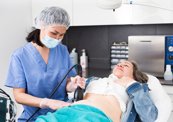 Qualified woman cosmetologist performing ultrasound cavitation body procedure for elderly female client at aesthetic cosmetology clinic.