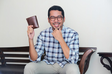 Adult Asian man sitting in the couch while smiling and showing his wallet