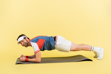 Positive sportsman standing in plank on fitness mat on yellow background.