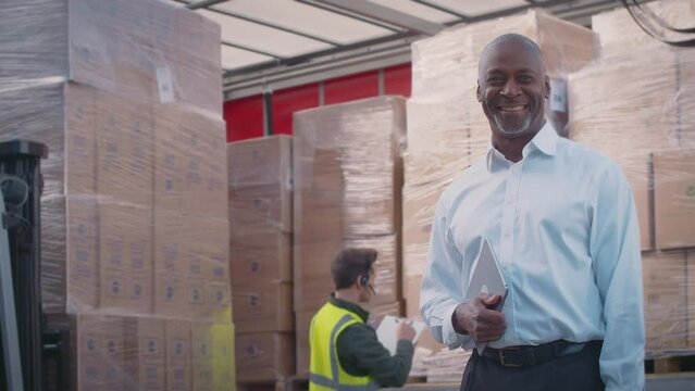 Smiling mature male freight haulage manager standing next to truck being loaded at distribution warehouse holding digital tablet looking into camera -shot in slow motion