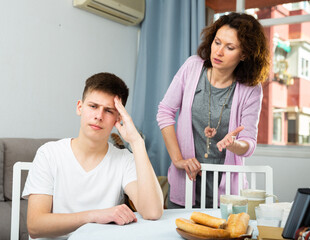 Portrait of troubled teen boy and his mother scolding him in home interior..