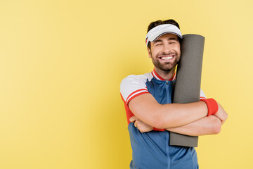 Cheerful sportsman in visor hugging fitness mat isolated on yellow.