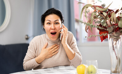 Fototapeta na wymiar Positive asian woman emotionally communicating with someone on her mobile phone, sitting at a table in an apartment