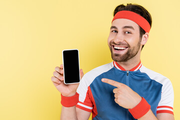 Positive sportsman pointing at smartphone with blank screen isolated on yellow.