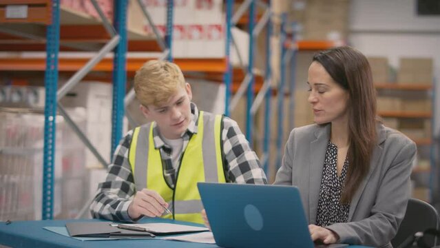 Female manager training male intern sitting at laptop in busy distribution warehouse - shot in slow motion