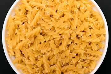 Uncooked Fusilli Pasta Lying on White Plate on Black Background. Raw and Dry Macaroni. Unhealthy and Fat Food - Top View