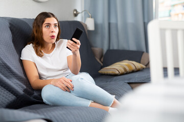 Dissatisfied young woman sitting on sofa with TV remote and watching boring movie at home