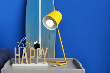 Table with stylish lamp and surfboard near blue wall in interior of living room