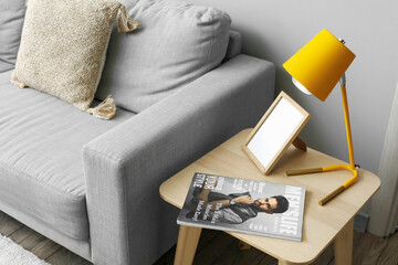 Table with stylish lamp, fashion magazine and photo frame near sofa in interior of living room