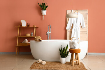Modern bathtub, houseplants, clean towels and robe near brown wall