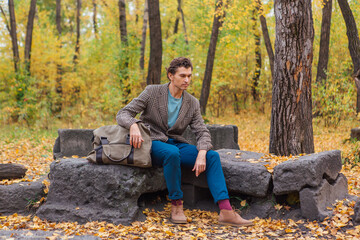 Fototapeta premium Tall handsome man dressed in a brown jacket posing with a bag on the rocks in autumn forest