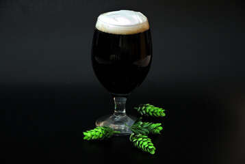 A tall glass of dark beer with dense white foam and hops on a black background.