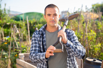 Young smiling man posing at smallholding with garden tools at sunny summer day