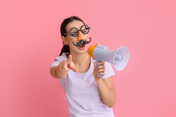 Young woman with funny disguise shouting into megaphone on pink background. April Fools Day celebration